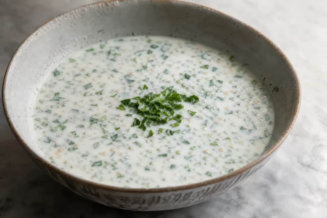 Creamy yogurt soup garnished with fresh herbs and wheat, served in a bowl.