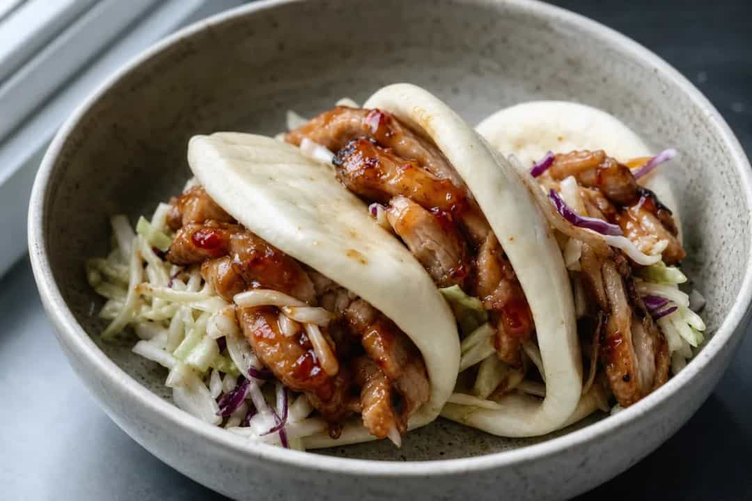 Plated bao sliders filled with sweet chili chicken, topped with vibrant crispy slaw and served on a wooden board.