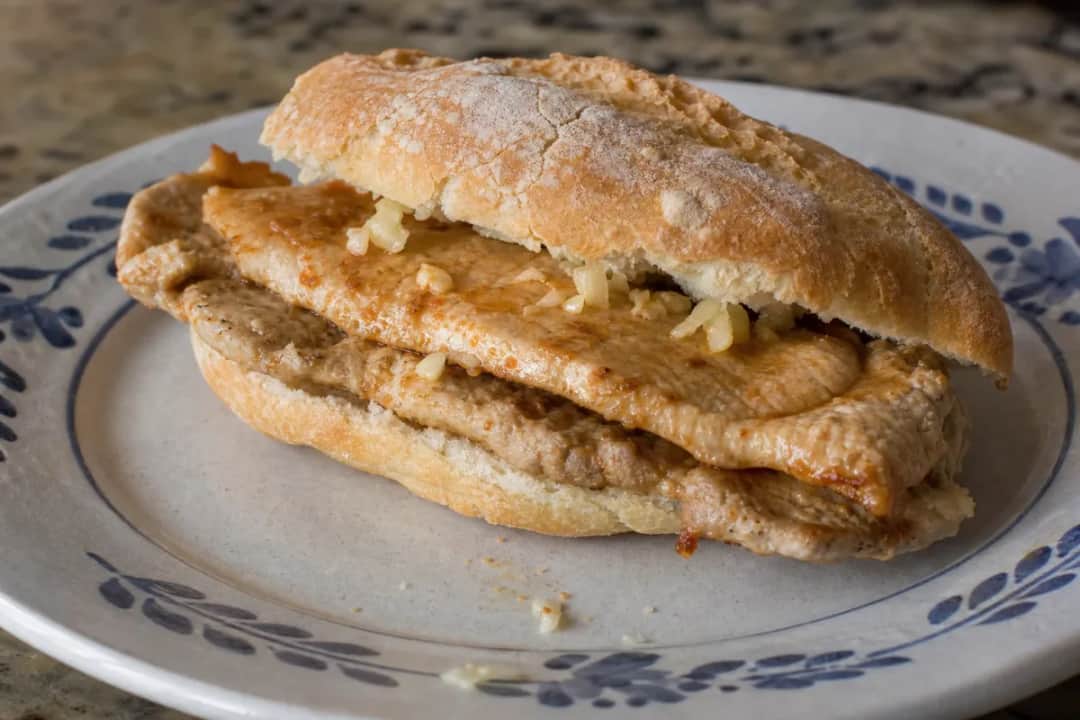 A hearty bifana sandwich featuring marinated pork slices in crusty bread, garnished with spices and served on a wooden board.