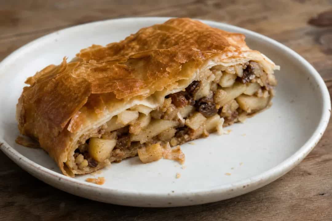Golden brown apple strudel with flaky crust, filled with spiced apples and nuts, garnished with powdered sugar.