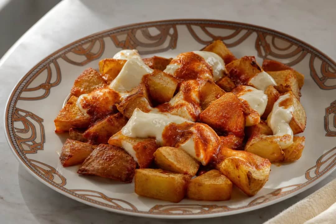 Crispy patatas bravas topped with spicy sauce and creamy aioli on a rustic plate.