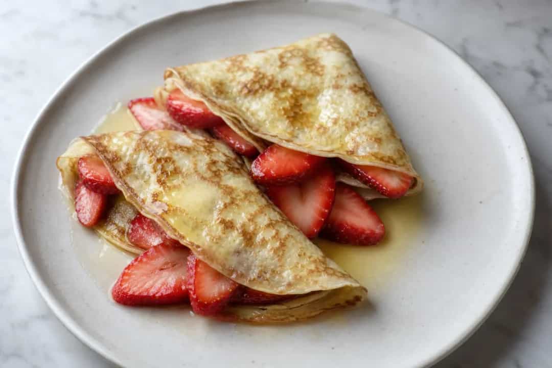 Thin crêpes folded and topped with fresh strawberries and whipped cream, garnished elegantly.