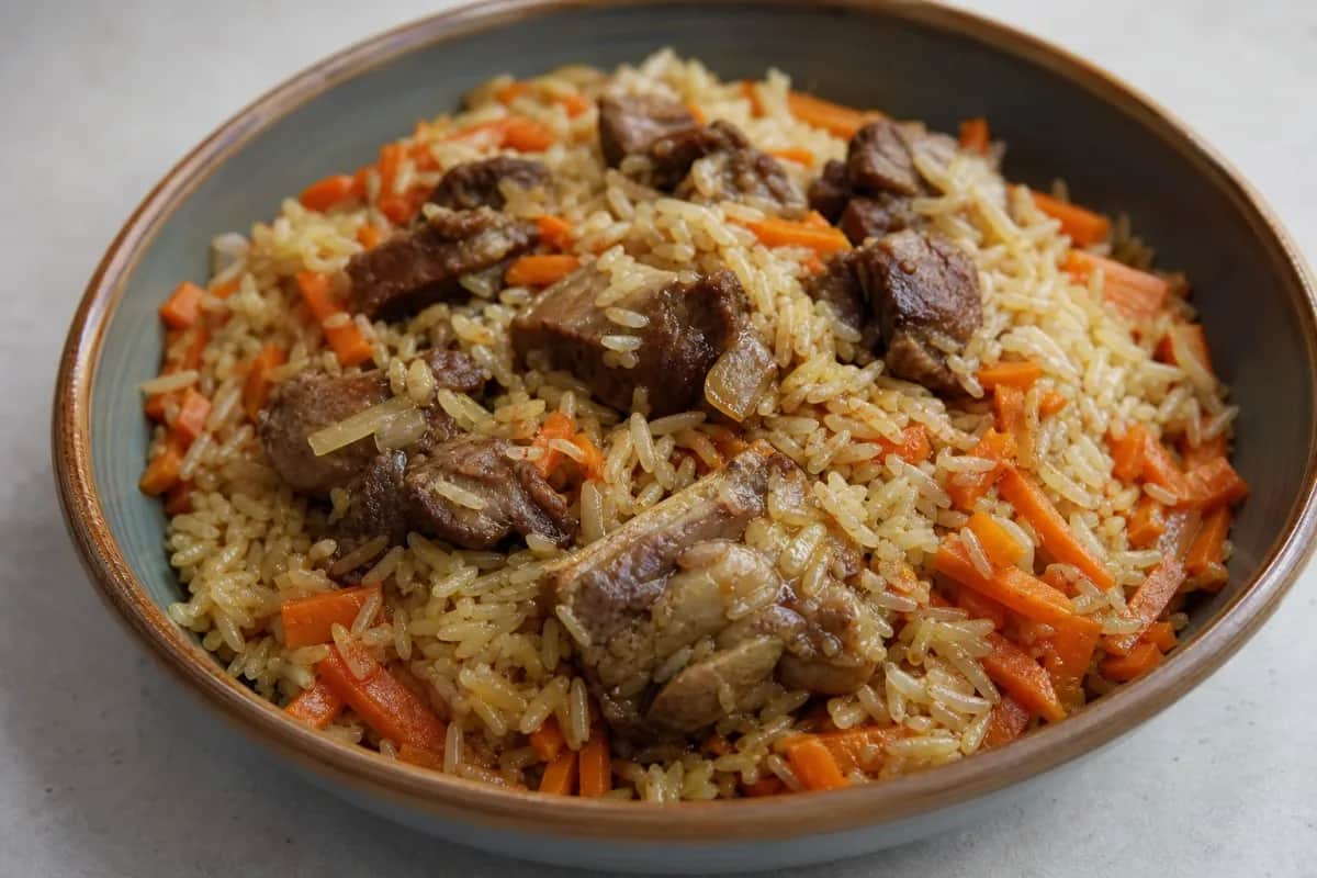 A plate of Kazakh plov featuring tender lamb, vibrant orange carrots, and fluffy rice garnished with herbs.