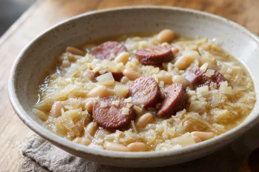 A bowl of hearty Jota stew with sauerkraut, beans, and smoked pork, garnished with fresh herbs.