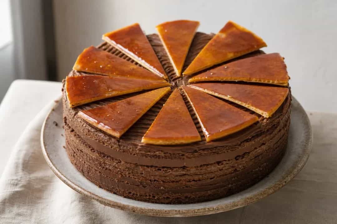 Layered Dobos Torte with glossy chocolate buttercream and caramel topping, garnished elegantly.