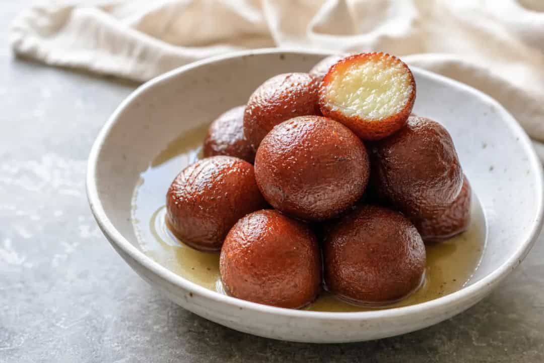 Soft, round gulab jamun soaked in golden syrup, garnished with pistachios and rose petals.