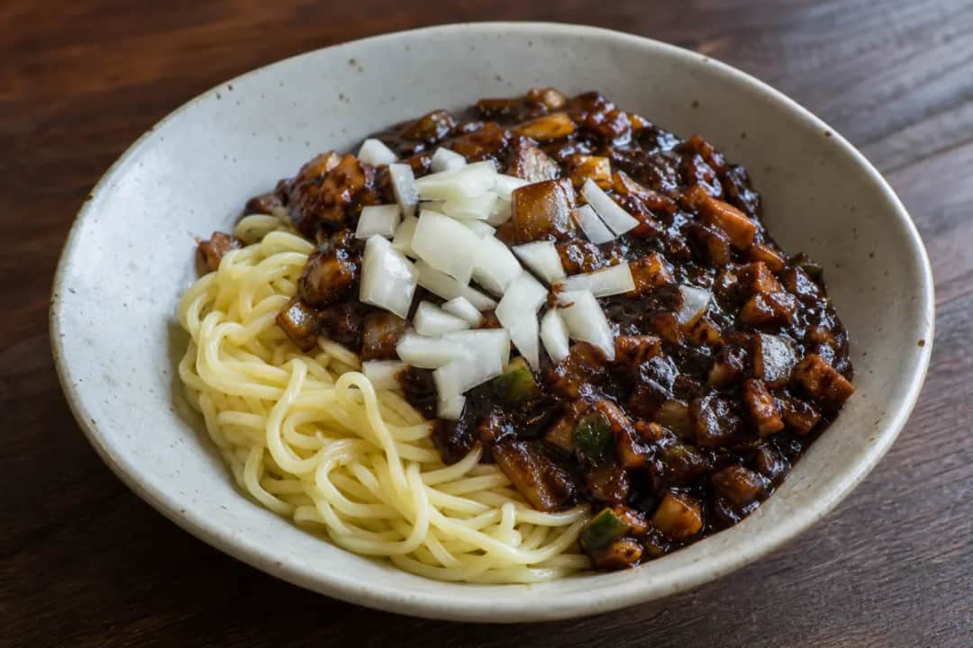 A bowl of Jajangmyeon featuring thick noodles covered in glossy black bean sauce, garnished with diced cucumbers and onions.