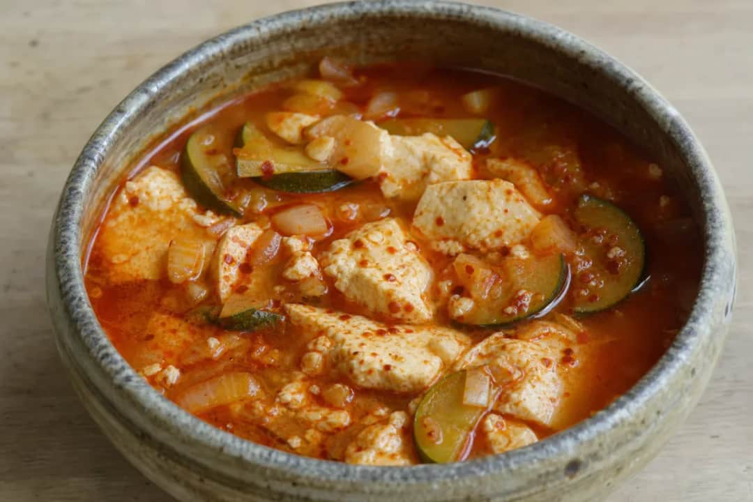 A bowl of Korean Sundubu Jjigae with soft tofu, vegetables, and a spicy broth, garnished with green onions.