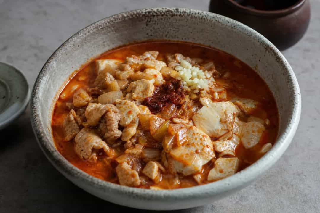 Bowl of Korean sundubu jjigae with soft tofu, vegetables, and spicy broth, garnished with green onions.