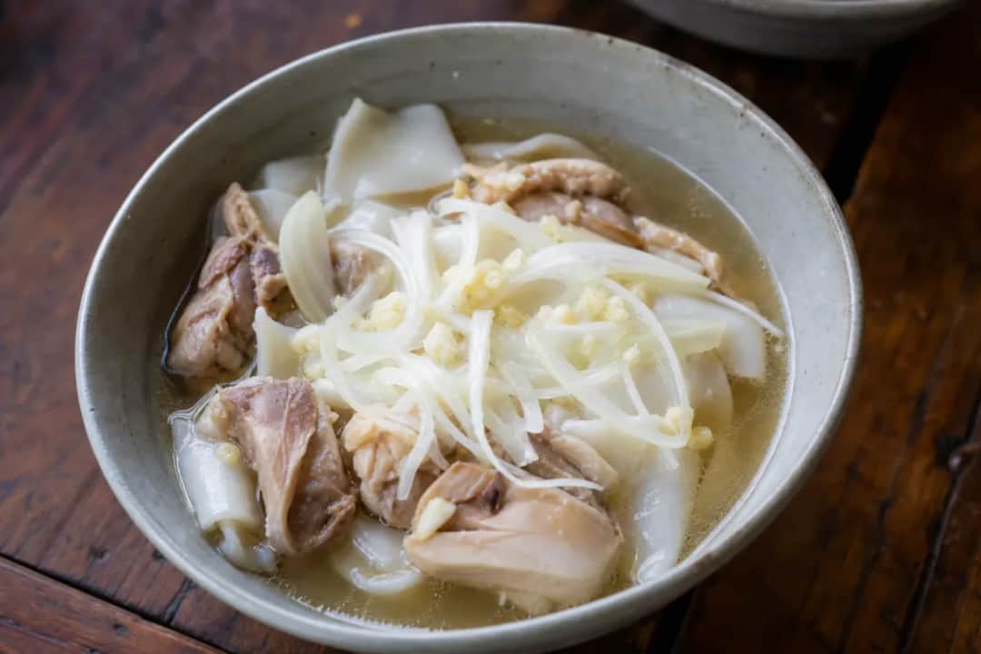 A bowl of Khao Piak Sen with smooth rice noodles, tender chicken, and fresh herbs garnished on top.