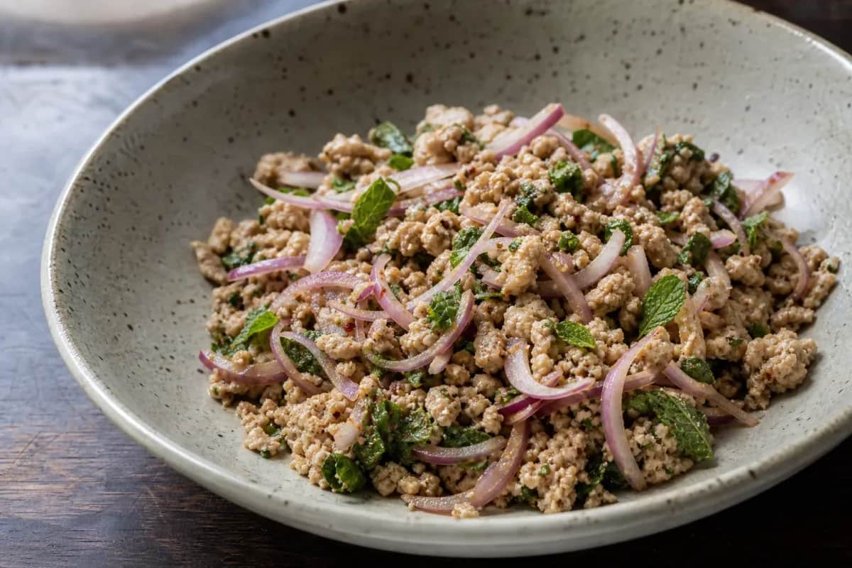 A colorful bowl of Laap featuring minced chicken, toasted rice, and fresh herbs garnished with lime.