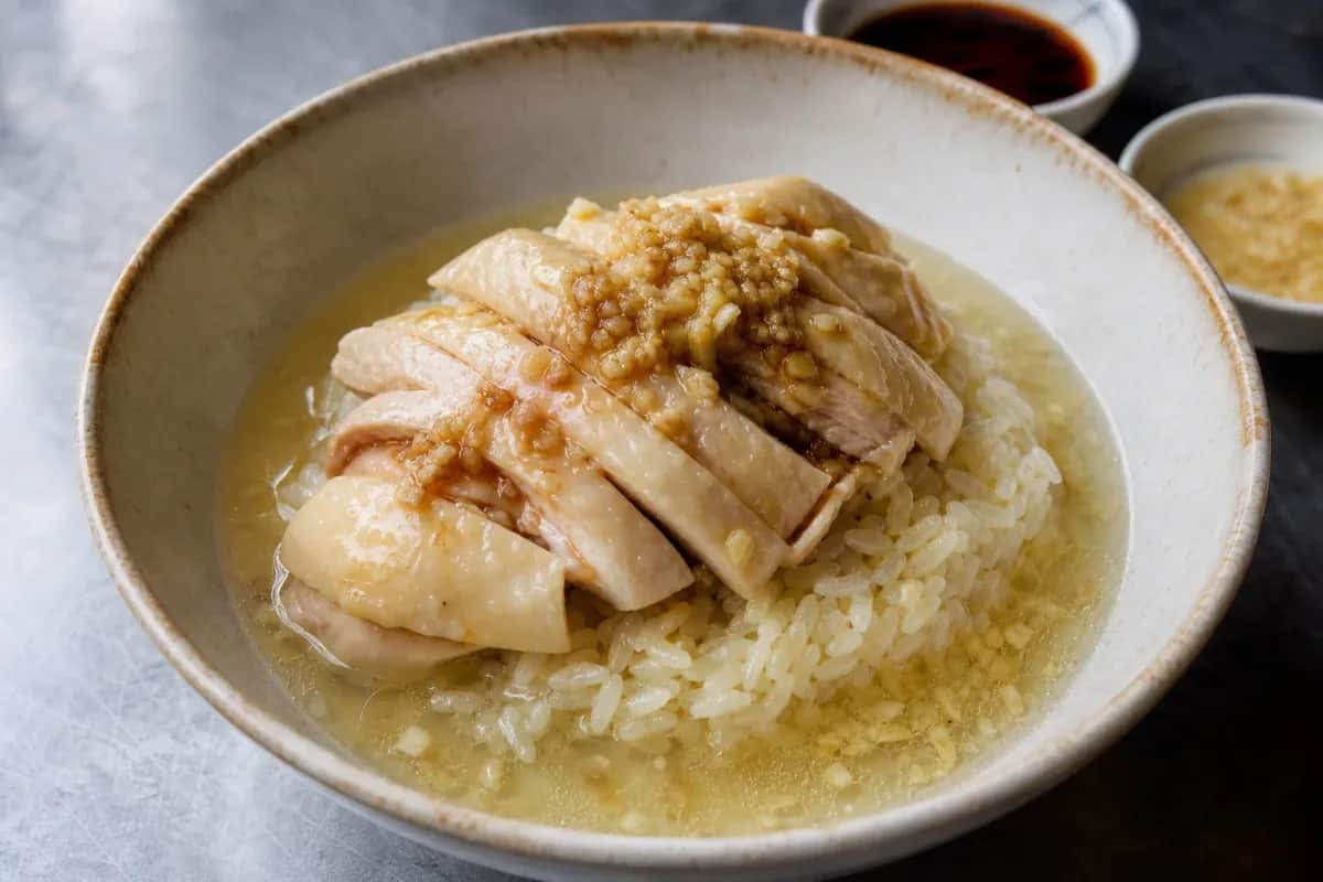 Plated Hainanese chicken rice with tender poached chicken, fragrant rice, and vibrant chili sauce garnished with herbs.