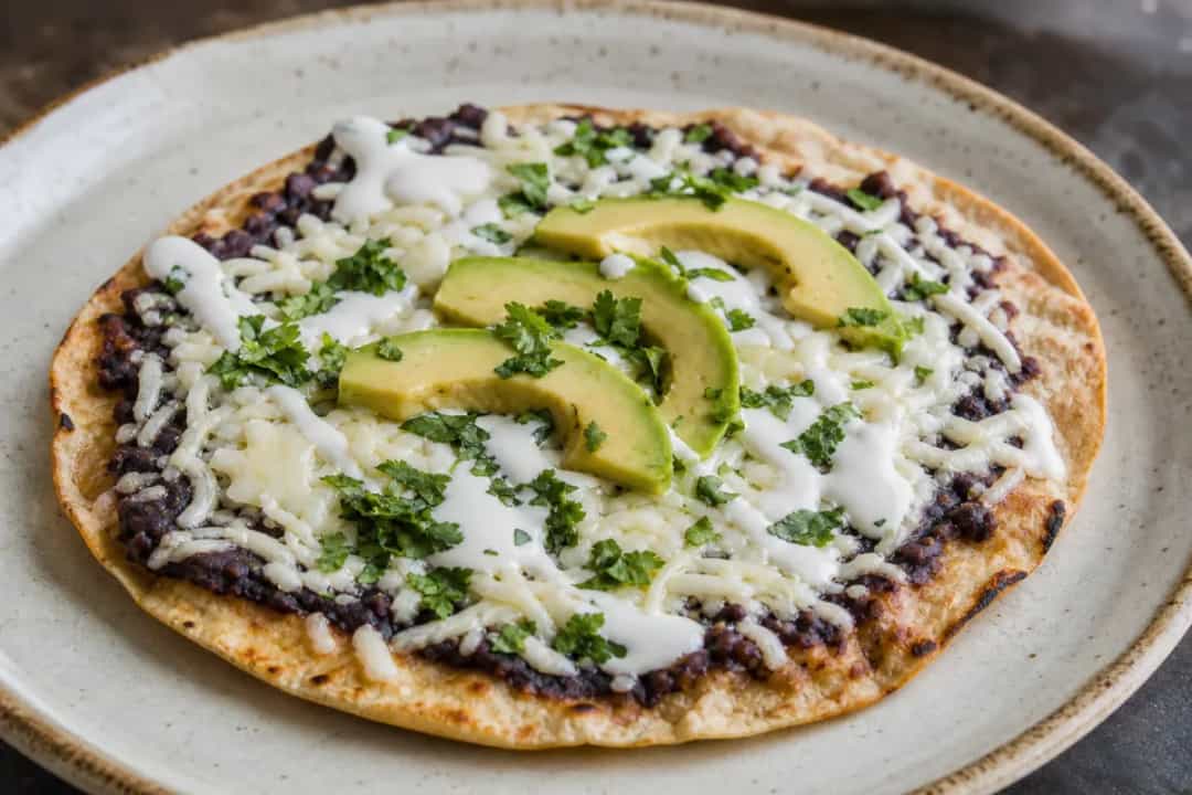 Crispy tlayuda topped with black beans, melted cheese, and sliced avocado, garnished with fresh cilantro.