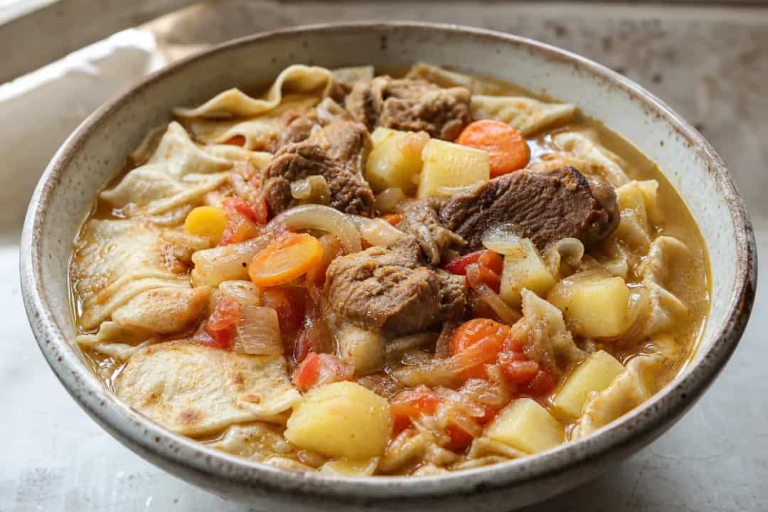 A hearty plate of thareed with tender lamb, vibrant vegetables, and flatbread, garnished with fresh herbs.
