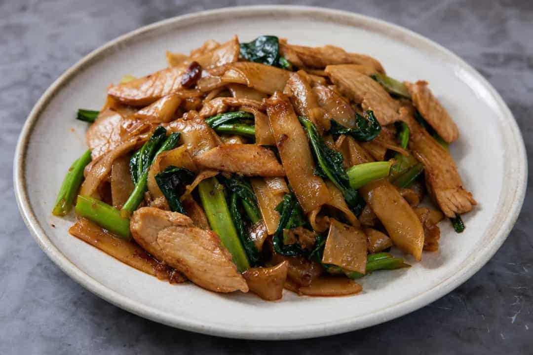 A colorful plate of Pad See Ew with chicken, Chinese broccoli, and wide noodles garnished with sesame seeds.