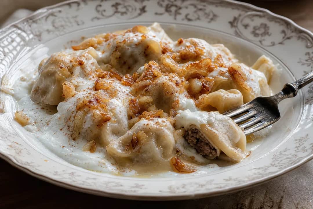 Plated Shish Barak dumplings in garlic yogurt sauce, garnished with crispy pine nuts.